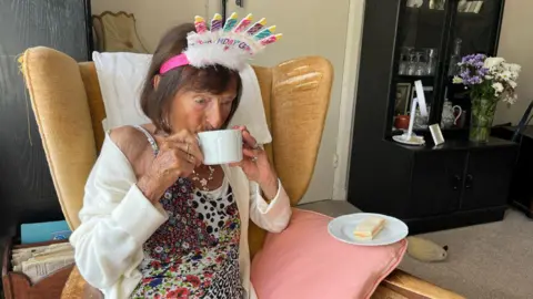 A woman wearing a white cardigan. She is drinking from a mug and sat in an arm chair.