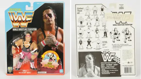 Vectis Auctions The Bret Hart figure in its packaging. The figure has a pink and black leotard on and has long black hair and is wearing his trademark pink sunglasses. The figure in encased ina plastic bubble on a car backing sporting a photo of Bret with the WWF logo. The back of the card is next to it and shows a variety of other figures available.