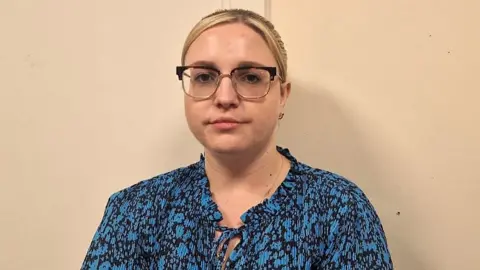 Sophie has blonde hair and wears glasses. She is wearing a blue blouse and looking at the camera. 