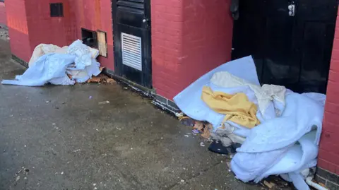 White bed sheets and a white duvet are in the rear doorway of a building, which has a red painted wall, black fire escape doors can be seen behind the bedding, there is a small pathway of concrete in front of the doors.
