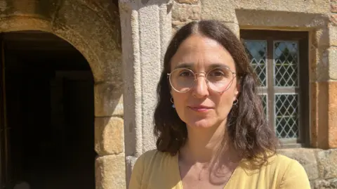 Annigna Kennedy wears round glasses and a light yellow top. She stands in bright sunlight in front of a stone building. An arched doorway is visible to the left, while a window with diamond-patterned panes is on the right. The textured stone walls and strong daylight create sharp shadows across the scene.
