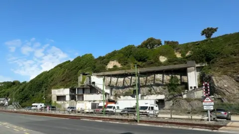A picture from across the road of the derelict site, you can see the remnants of a building on the cliff side and some cars parked in front.