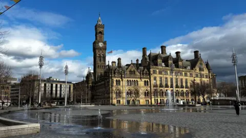 A large Gothic-style civic building with a tall clock tower overlooks a wide public square with patterned paving and water fountains.