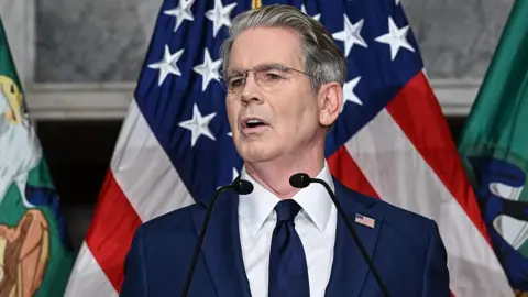 US Treasury Secretary Scott Bessent speaks during a press conference at the Cash Room of the Treasury Department, as he announces a new set of sanctions against Iran. He is wearing a dark blue suit with a white shirt and dark blue tie. He has grey hair and wears glasses.