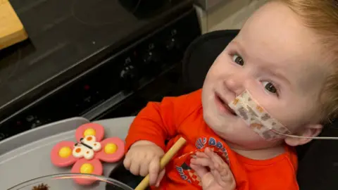 Kasey Mumford A young boy with ginger hair is wearing a red long sleeved top and is holding a wooden spoon and a bowl filled with chocolate. A tube is coming from his nose with a plaster over the top. 