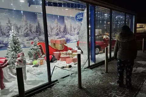 MG Accident Repair Centre The photo is from outside the showroom looking into the display after the theft. Broken glass is all over the floor and the display of presents and the Grinch scattered about. A sleight and a red car can also been seen in the image.