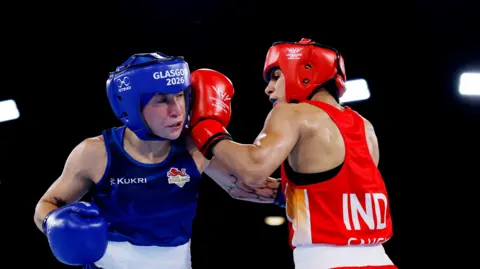 Ruby White and Sakshi Chaudhary wearing protective headgear exchange punches in a boxing ring. White wearing blue representing England is struck by an Chaudhary in red representing India under bright arena lights.