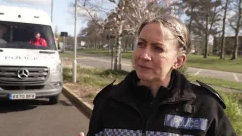 A female police officer with blonde hair tied back. She is wearing a black police uniform with blue and silver patches. One says "police".
There is white police van behind her.