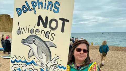 Surfers Against Sewage A woman holding a sign saying "dolphins not dividends" on a beach