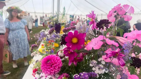 Cheryl Dennis/BBC A man and a woman stand inside a large white marquee looking at a large floral display of pink and dark purple flowers, which is laid out on a long white table