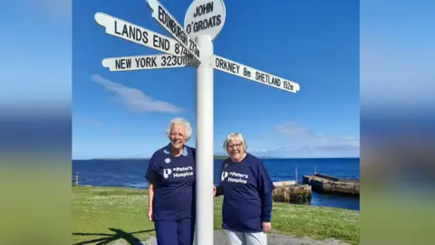 Valerie and Sue are standing next to a white direction sign post in John O'Groats. The post is pointing in multiple directions to New York, Edinburgh and Lands End.  