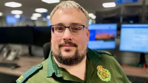 Mark Norman/BBC A man with a beard and glasses in green ambulance service uniform sitting in an ambulance 999 call centre with computer screens in the background.