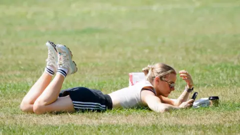 A woman wearing a T-shirt, shorts, trainers and sunglasses lies on a patch of grass looking at her phone. 