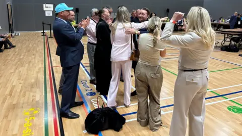People dressed smartly hug each other and clap while looking happy in a sports hall at an election count