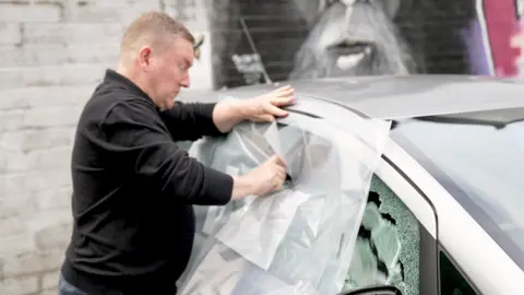 Steve Hay holds protective film to a car window as he breaks it with a tool. There is a graffiti-covered brick wall in the background.