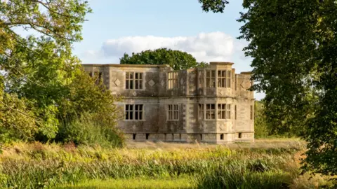 National Trust Images/Mike Selby A view of Lyveden Lodge through trees and across grass. It shows a roofless late Elizabethan stone built building. 