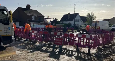 Plastic barriers form a square on a flooded road, and a sign reads "footpath closed"