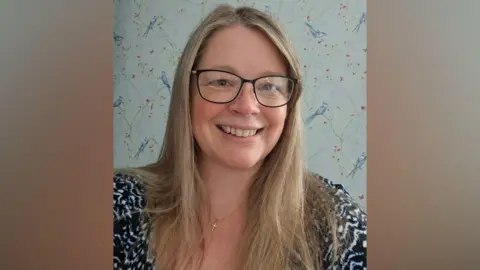 Supplied A woman wearing black-framed glasses, wearing a patterned blouse. She has blonde hair and is smiling at the camera.