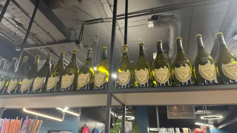 Alice Cunningham/BBC Bottles of Dom Perignon champagne bottles rest of a shelf. Each one details deals Halo has successfully set up with high profile companies.