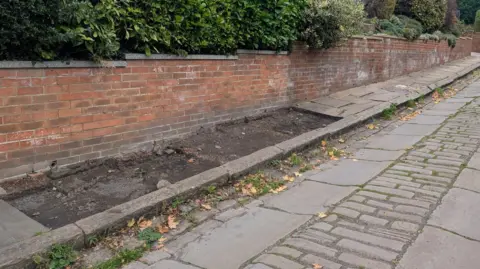 Alex Moss/BBC A residential street with A cobblestone and stone-slab pavement running along the right side. Several large slabs are missing from the pavement, leaving bare patches of earth and uneven gaps. 
