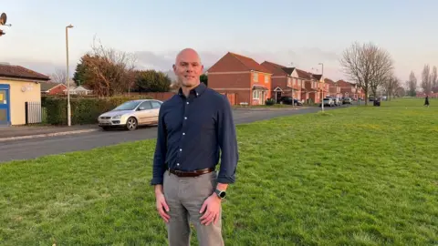 A bald middle aged man who is wearing a navy, long-sleeved shirt and grey trousers stands on a green space bordered by houses and a residential street in a deprived area of Gloucester