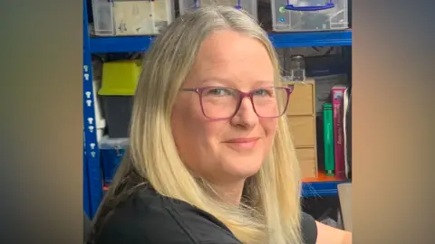 Prickly Pigs Hedgehog Rescue Portrait photograph of a person with long, light-coloured hair wearing pink rectangular glasses and a dark top, seated in front of blue metal shelving. The shelves contain clear plastic storage boxes, wooden drawers, books, jars and assorted workshop or office supplies. The person is turned slightly towards the camera, with the face in sharp focus and the background softly blurred.