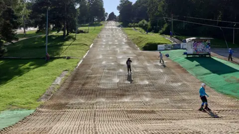 Gloucester Ski and Snowboarding Centre A view from the bottom of the slope. There are several skiers and a snowboarder on the slope and a ski lift on the right. It is a sunny day and trees line either side of the slope.
