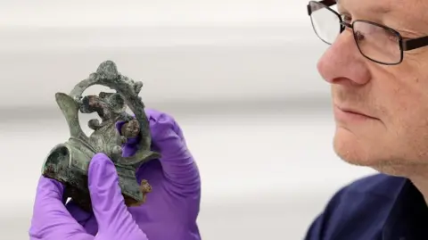 A man wearing glasses and a rubber gloves holds a decorative copper alloy finial that's been cleaned up 