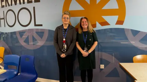 Two women stand in front of a large image on a wall, where the word school can be seen, as well as a repeated logo. Both have their hands crossed in front of them and are smiling.