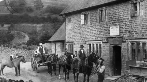 CCBS A black and white photo of the pub in the 1900s, with a sign above the door lintel saying "New Inn". There are several horses outside, one with a cart behind, and several men and boys in work clothes.