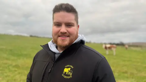 A man, lasting  successful  beforehand   of a tract  of grass, looking astatine  the camera. He is pictured from the thorax  up. He is wearing a grey hoodie and a achromatic  overgarment   with a yellowish  logo with a cattle  connected  it. He has abbreviated  brownish  hairsbreadth  and a choky  beard. There is simply a cattle  successful  the inheritance  to the close    of the man. The entity  is afloat  of grey clouds. 