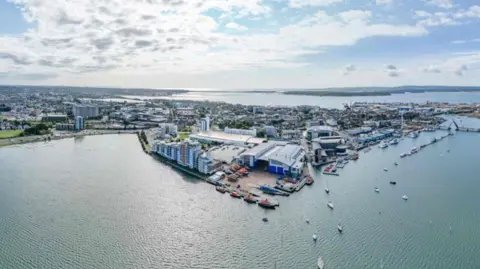 Aerial view of Poole town and waterfront with the RNLI College in the foreground and Twin Sails Bridge on the right. The outline of Brownsea Island and Sandbanks can be seen in the distance on the horizon.