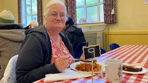 Shariqua Ahmed / BBC Angie wearing a black hoodie and a red dress and holding a knife and fork smiling for the camera