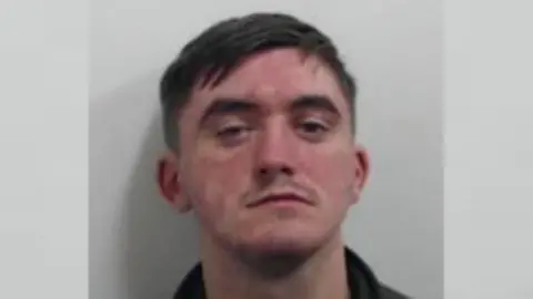 Police Scotland A mugshot of a man with brown hair and a faint moustache. He is wearing a black top.