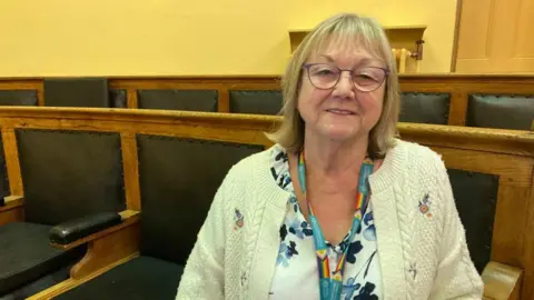 A woman sitting in a council chamber. She is wearing glasses and smiling at the camera.