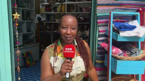 A woman with her hair in braids stands in front of a shop - she is holding a microphone and looking into the camera.