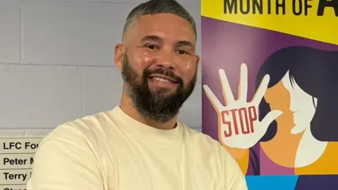 Tony Bellew, 43, who has dark hair speckled with grey and a large black beard, smiles at the camera in front of a sign with slogans including 'Schools Month of Action' and a cartoon image of a woman holding up her hand with the word 'Stop' written on her palm in red lettering. 