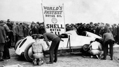 Getty Images An archive picture of 'Babs' with mechanics at each wheel. There is a group of spectators behind the car watching. A sign behind the vehicle says 'Fastest motor oil Shell motor' with the Shell logo. 