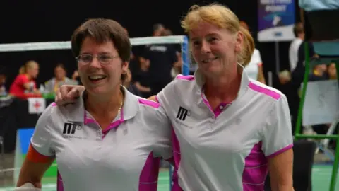 Pamela Peard The two ladies stand arm in arm. Pam on the left with short brown hair smiling, wearing thin lensed glasses. Right is Sian, also smiling, wearing her blonde hair in a ponytail. The two wear white polo shirts, with bright pink accents.