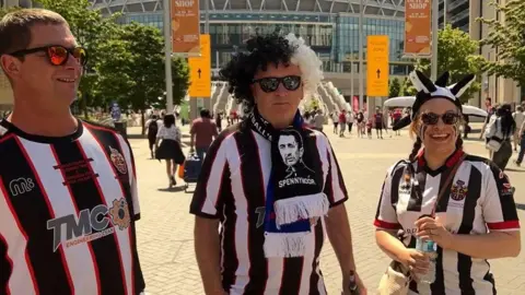 Spennymoor fans Sean, Alan and Claire on Wembley Way hours before kick-off. They're all wearing  black, white and red football kit tops. Alan also has a black and white Spennymoor Town scarf folded around his neck and has one a black and white fancy dress curly hair wig. Claire is wearing a black and white jester's hat.  It's sunny and they're all wearing sunglasses.
