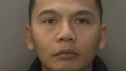 Le has close cropped hair in his police mugshot. He is wearing a dark top.