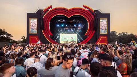 A large crowd of people gathered in front of the heart shaped stage at Love Saves the Day festival. It is dusk and the sky is a very pale orange. 