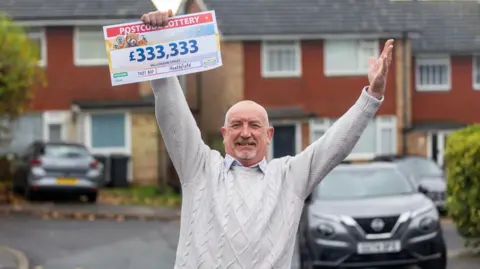 People's Postcode Lottery A mature man wearing a white jumper smiling to camera standing in a cul de sac with cars and houses behind him holding up a giant cheque