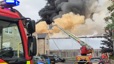 Black, orange and grey smoke rises in a huge plume from a large grey industrial building which sits behind a wooden fence at the end of a residential street. The houses are made of larger beige stone bricks and sit over two storeys. They are all of similar design like so many private housing estates. Lots of cars are parked in front of the houses. The cab of a fire engine is in the foreground. In front of it, a flat bed fire vehicle has a ladder mounted on it. It is extended towards the flames with, presumably, firefighters directing water towards the flames from the cherry picker basket at the end of the ladder. Two firefighters in hi-vis and helmets stand in the distance, backs to shot.