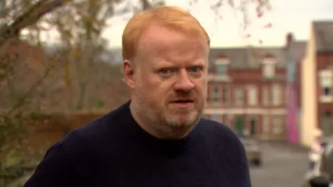 A red haired man with a red and white beard is wearing a navy top and looking to the camera. Behind him out of focus is a street with red brick houses and cars. A tree can also be seen behind him. 