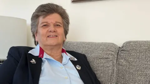 Solange sits on a light gray sofa indoors, wearing a light blue shirt with a dark blazer featuring decorative pins on the lapels. A lamp and a framed black-and-white tree photograph are visible in the background.