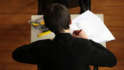 A view of a student sitting an exam. They are seen from behind filling in an exam booklet with their answers. There is also a clear plastic folder containing pens and pencils.