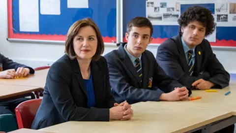 PA Media Bridget Phillipson sitting in a classroom next to two boys. She is leaning on a desk.