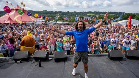 PA Media Joe Wicks on stage in a blue top, grey shorts and white trainers looking at the camera with his arms in the air. He is stood on a stage with a large crowd behind him.