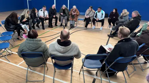 BBC/Stuart Whincup Seventeen people sat in a circle in a sports hall on blue chairs. One of them is a man in what appears to be police uniform.
